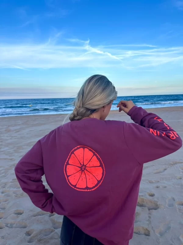 chica en la playa con un pantalón campana gris oscuro y una sudadera de pink pomelo en color burdeos con un estampado grande en espalda de una rodaja de pomelo en color coral flúor