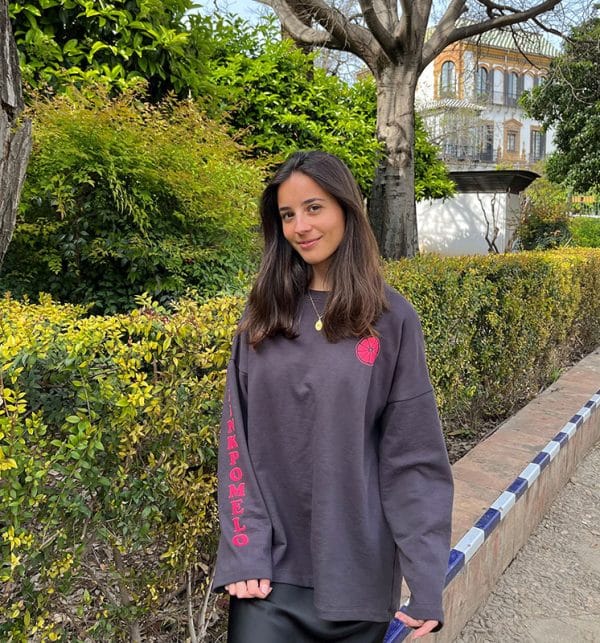 chica en una calle con jardín con una sudadera oversize gris antracita con print en pecho izquierdo de una rodaja de pomelo en coral flúor y texto pink pomelo en manga derecha en coral flúor