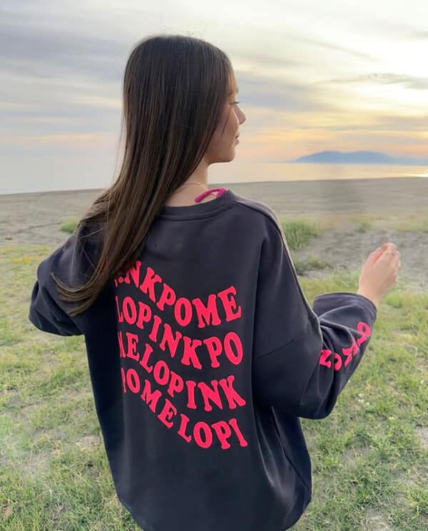 chica en la playa con una sudadera oversize en gris antracita con texto pinkpomelo en ondas en color coral flúor en espalda y manga derecha