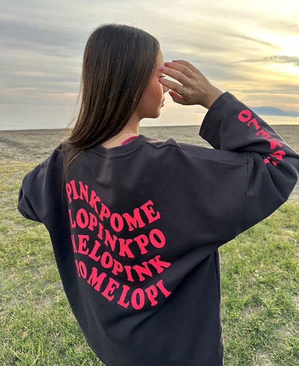 chica en la playa con una sudadera oversize en gris antracita con texto pinkpomelo en ondas en color coral flúor en espalda y manga derecha