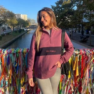 chica sonriendo delante de un puente con cintas de colores con una sudadera de media cremallera buganvilla con pieza central en marino con texto bordado pink pomelo y bordado en manga de rodaja pomelo en marino