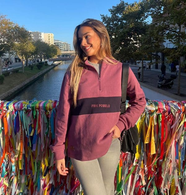 chica sonriendo delante de un puente con cintas de colores con una sudadera de media cremallera buganvilla con pieza central en marino con texto bordado pink pomelo y bordado en manga de rodaja pomelo en marino