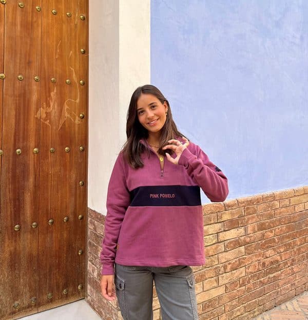 chica sonriendo delante de una puerta de madera con una sudadera de media cremallera buganvilla con pieza central en marino con texto bordado pink pomelo y bordado en manga de rodaja pomelo en marino