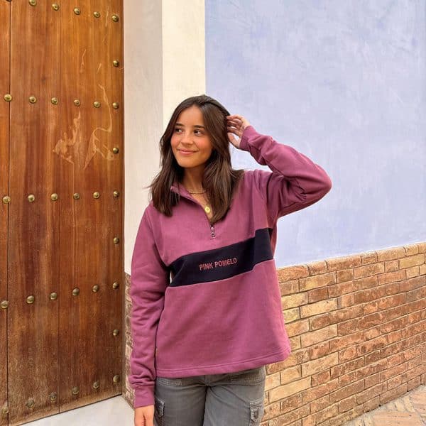 chica sonriendo delante de una puerta de madera con una sudadera de media cremallera buganvilla con pieza central en marino con texto bordado pink pomelo y bordado en manga de rodaja pomelo en marino