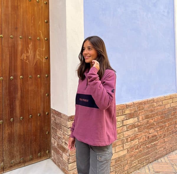 chica sonriendo delante de una puerta de madera con una sudadera de media cremallera buganvilla con pieza central en marino con texto bordado pink pomelo y bordado en manga de rodaja pomelo en marino