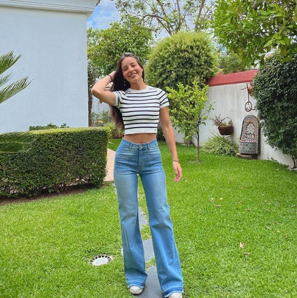 chica en un jardín con una mano en el pelo con un top blanco de rayas azul marino manga corte y tejano azul claro