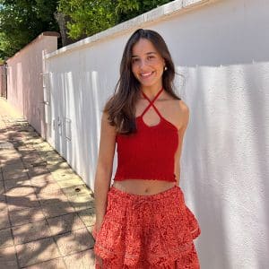 chica sonriendo en la calle delante de una pared blanca de una casa con un top rojo fruncido y atado al cuello y falda corta con dos volantes coral y naranja