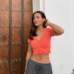 chica delante de una puerta de madera con un pantalón fluido gris antracita cargo y top naranja corto naranja con cordón frontal para fruncir