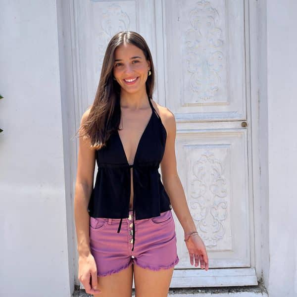 chica posando delante de una puerta blanca con un short denim rosa desflecado en bajo y cierre con botones plateados y un top negro anudado al cuello y escote pico