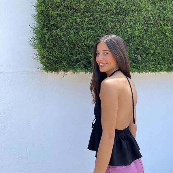 chica posando en la calle con un short denim rosa y un top negro anudado al cuello y espalda al aire, solo tiene una pieza en el bajo de la espada ajustada con goma elástica