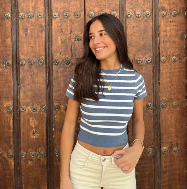 chica sonriendo delante de una puerta de madera con un top de punto de rayas azules y blancas y con un pantalón beige