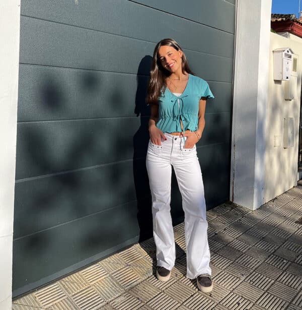 chica morena delante de un portón de garaje color gris con un top verde abierto en frontal y cerrado con 2 cordones en manga corta y pantalón blanco con tachuelas plateadas