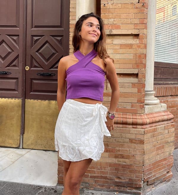 chica apoyada en la pared con un top morado corto cruzado al cuello y falda corta blanca