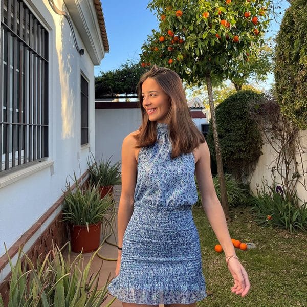 chica sonriendo en un jardín con vestido corto con gasa estampada azul con cuello halter y espalda descubierta con la parte de la falda fruncida y forro interior