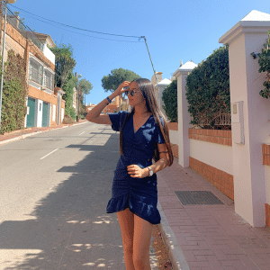 chica tocándose el pelo con gafas de sol y con un vestido corto azul marino fruncido en el centro y volante en bajo