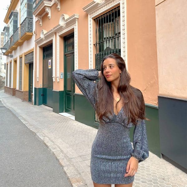 chica tocándose el pelo con una vestido de fiesta en tonos plata de manga larga en forma murciélago, escote en pico y la parte de la falda ajustada.