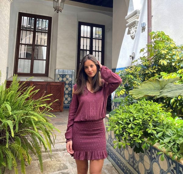 chica en un patio andaluz lleno de plantas verdes con un vestido fruncido en su parte de falda y hombros descubiertos en color granate con lúrex dorado en forma de círculos