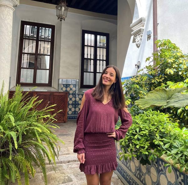 chica en un patio andaluz lleno de plantas verdes con un vestido fruncido en su parte de falda y hombros descubiertos en color granate con lúrex dorado en forma de círculos