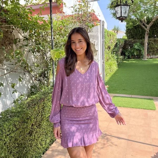 chica sonriendo en un jardín con un vestido rosa viejo con lúrex plateado de manga larga, hombros descubiertos, parte de la falda con fruncido y volante en bajo