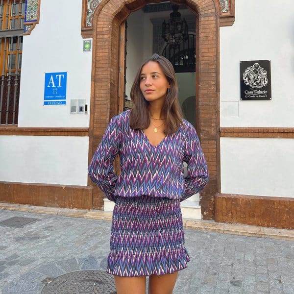 chica en la calle delante de una puerta abierta con un vestido print zigzag en colores negros y lilas con escote pico y falda fruncida con gomas