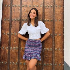 chica sonriendo apoyada en una puerta de madera con una blusa blanca y falda corta zigzag